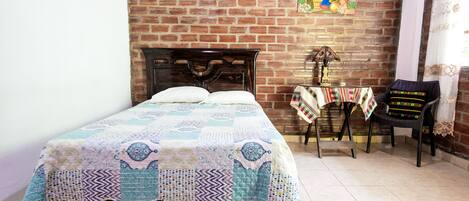 A room with a brick wall, a bed with a quilt, a small table with a lamp, and a chair with a colorful cushion.