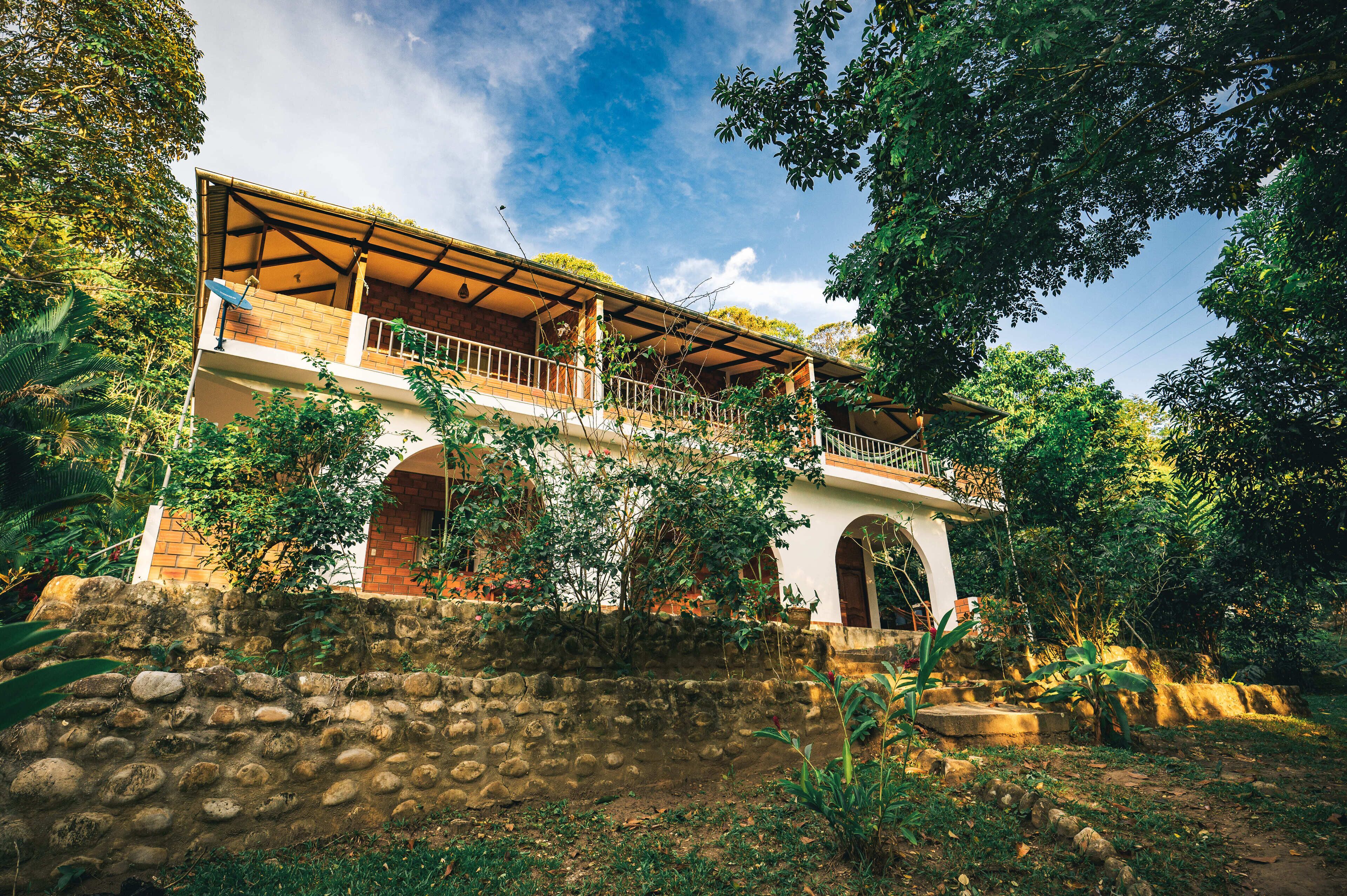 A two-story house with a balcony, surrounded by lush greenery and a stone wall.