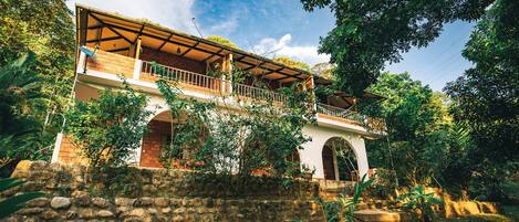 A two-story house with a balcony, surrounded by lush greenery and a stone wall.