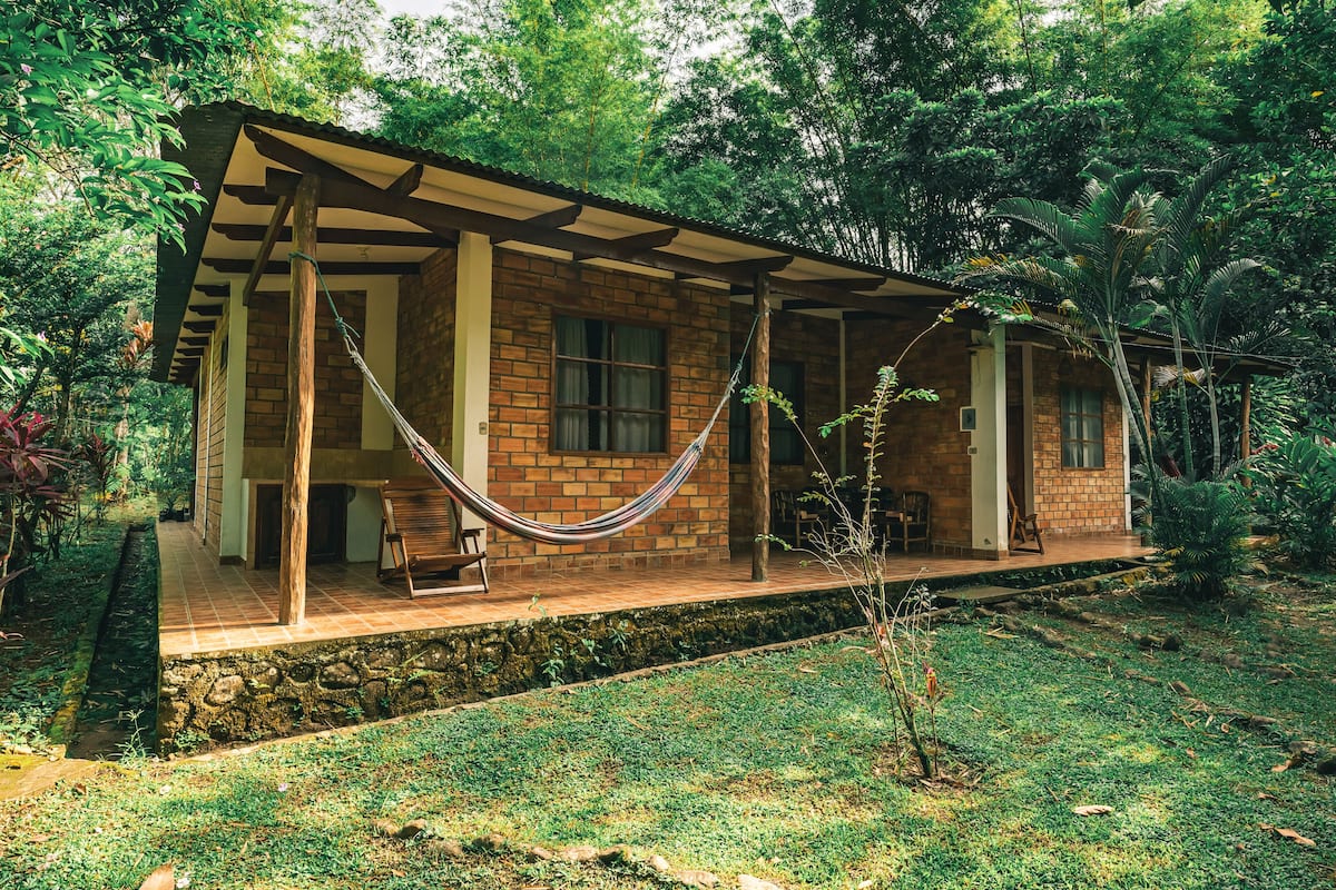 Uma casa rústica com varanda, uma rede e um caminho de pedra.