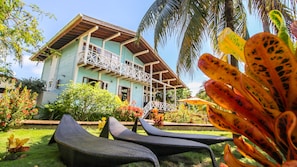 Una casa de dos pisos con balcón, rodeada de exuberante follaje y plantas tropicales.