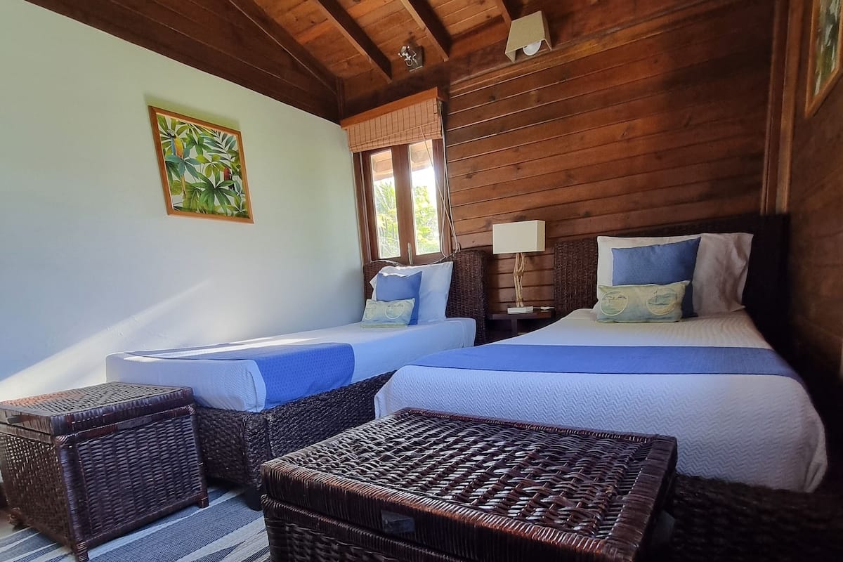 A room with two beds, wicker furniture, a striped rug, and a painting on the wall.
