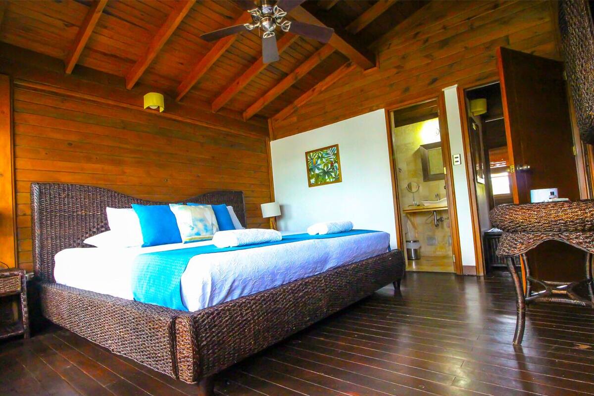 A wooden-paneled bedroom with a large bed, a ceiling fan, and a bathroom visible through an open door.
