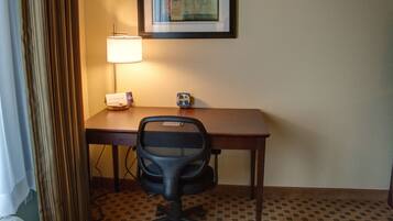 A hotel room with a desk, chair, lamp, and a framed abstract painting on the wall.