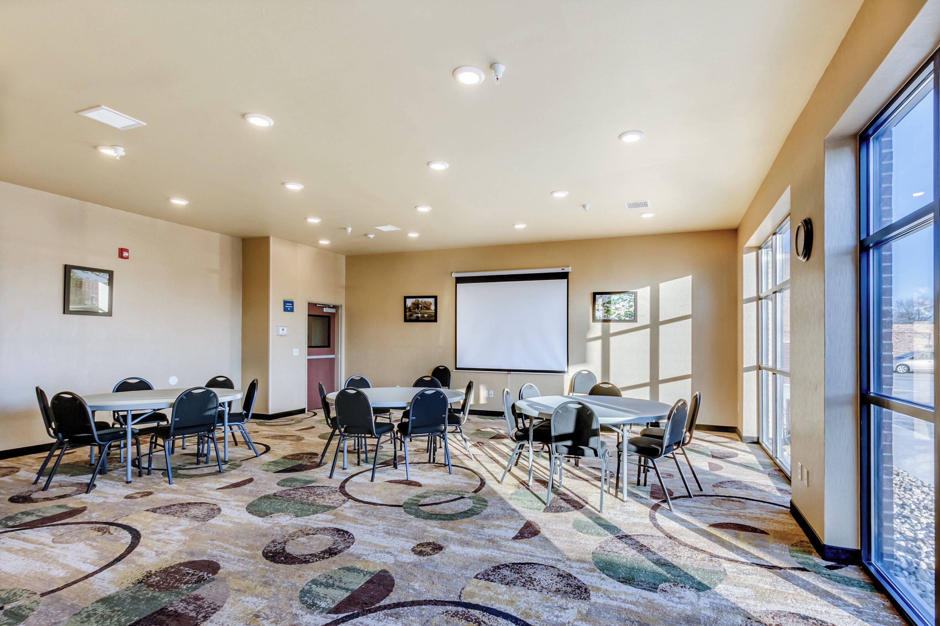 Une salle de conférence équipée de plusieurs tables et chaises, d’un écran de projection et de grandes fenêtres.