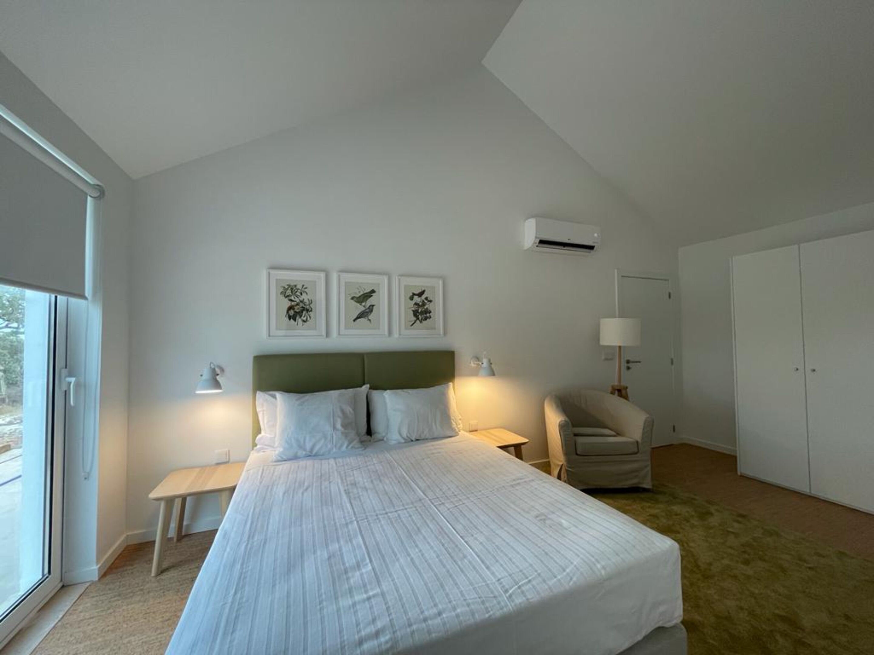 Un dormitorio moderno con una cama grande, una mesita de noche, una silla y un armario. Hay tres cuadros enmarcados en la pared y un equipo de aire acondicionado sobre la cama.