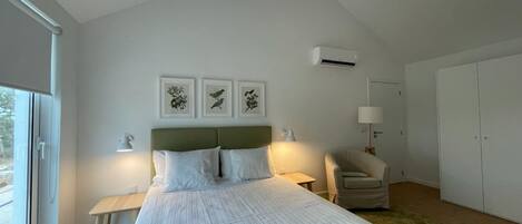 Un dormitorio moderno con una cama grande, una mesita de noche, una silla y un armario. Hay tres cuadros enmarcados en la pared y un equipo de aire acondicionado sobre la cama.