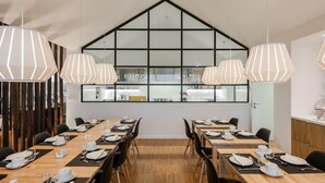 Un comedor moderno con mesas largas dispuestas para una comida, ventanas amplias con un diseño geométrico e iluminación colgante.