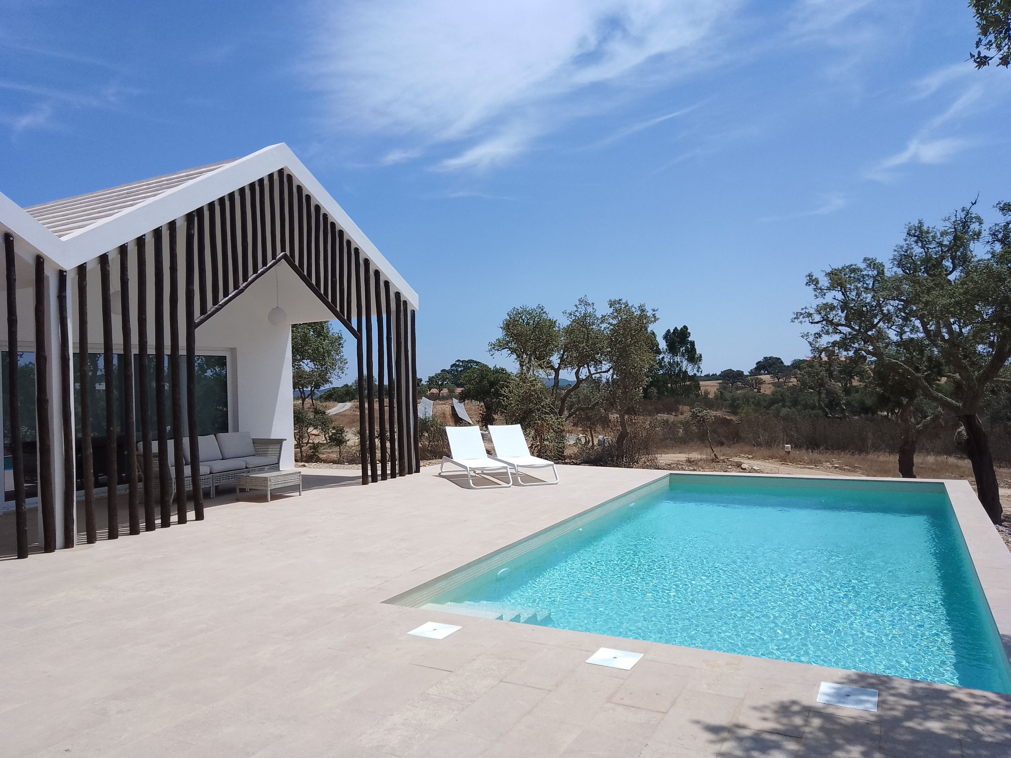 A modern outdoor pool area with a clear blue pool, a covered seating area with lounge chairs, and a view of the surrounding landscape.