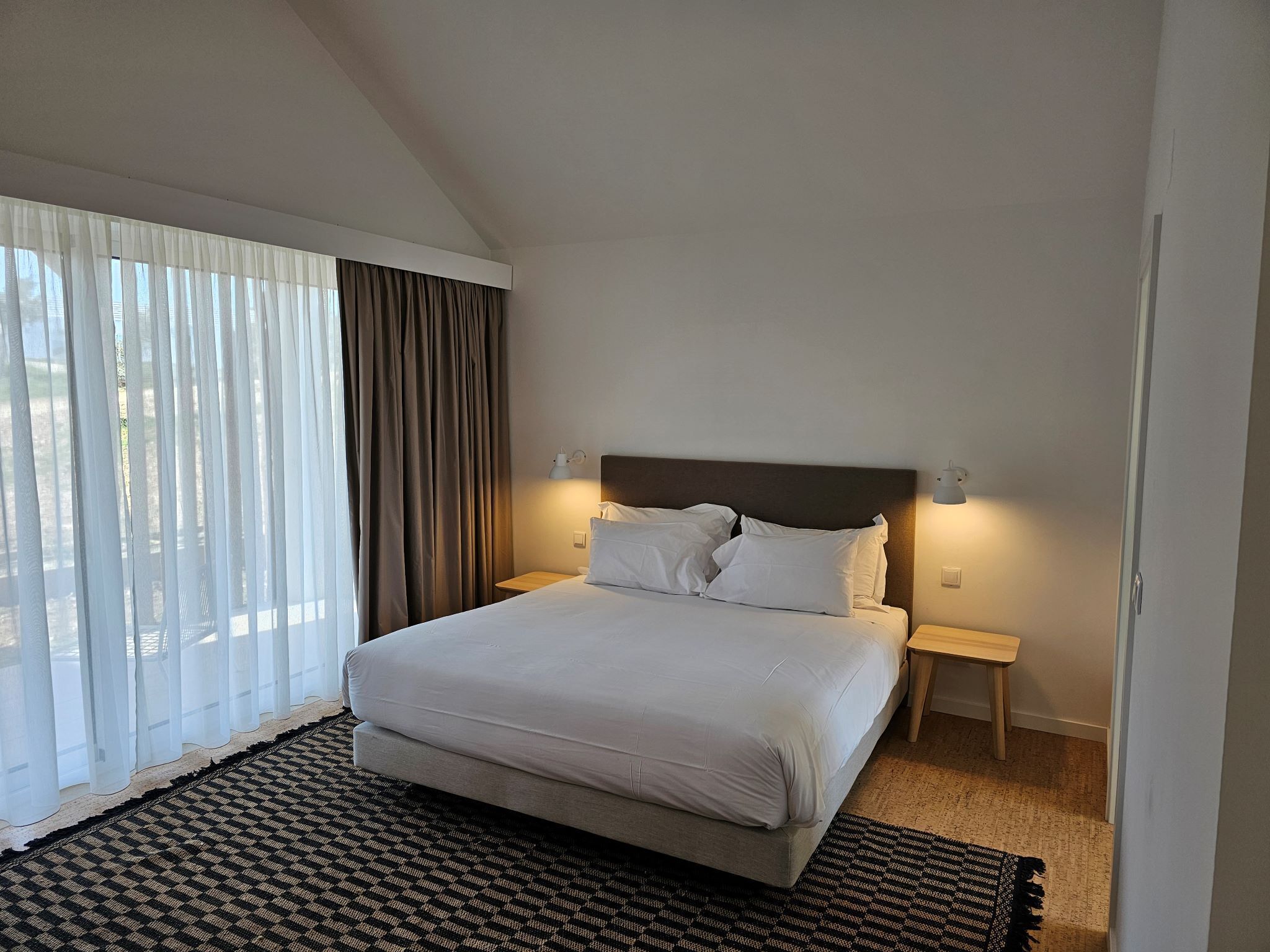 A hotel room with a bed, two bedside tables, a headboard, and a large window with curtains.