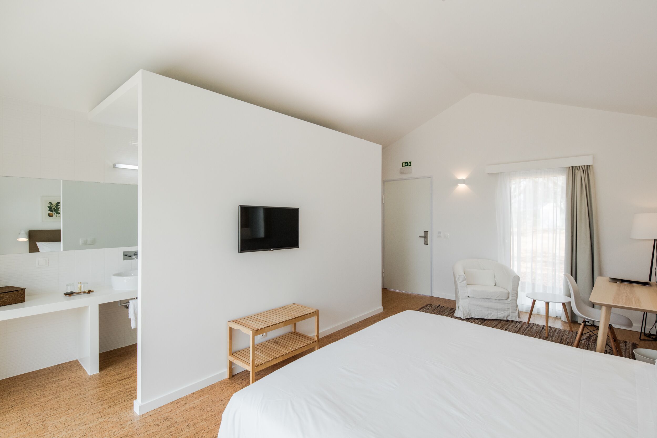 A modern hotel room with a large bed, a desk, a chair, a TV, and a bathroom with a mirror and sink.