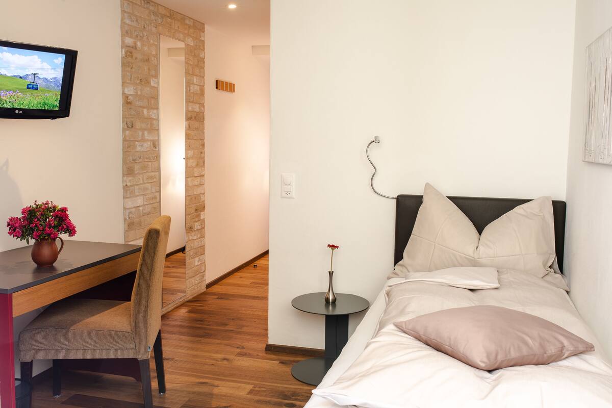 Moderni hotellihuone, jossa on puulattia, valkoisilla tekstiileillä varustettu sänky, pieni pyöreä pöytä maljakolla, tuoli ja seinälle asennettu televisio.