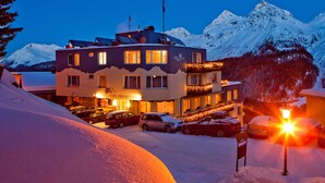 A hotel with a snow-covered mountain backdrop, multiple cars parked in front, and a lit sign indicating the hotel's name. - Hotel Hohe Promenade Arosa (Arosa)