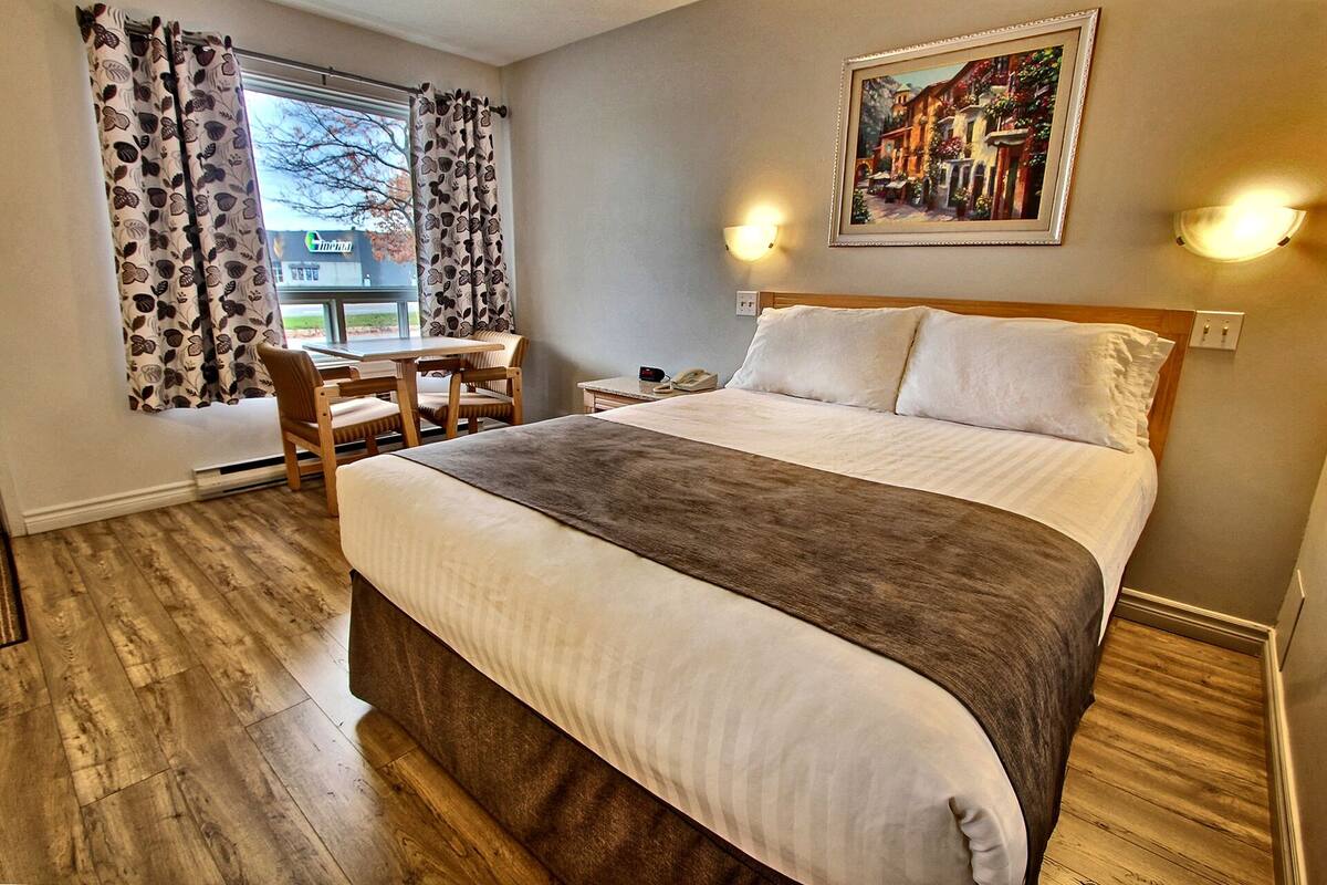 A hotel room with a large bed, a desk with a chair, a painting on the wall, and a window with curtains.