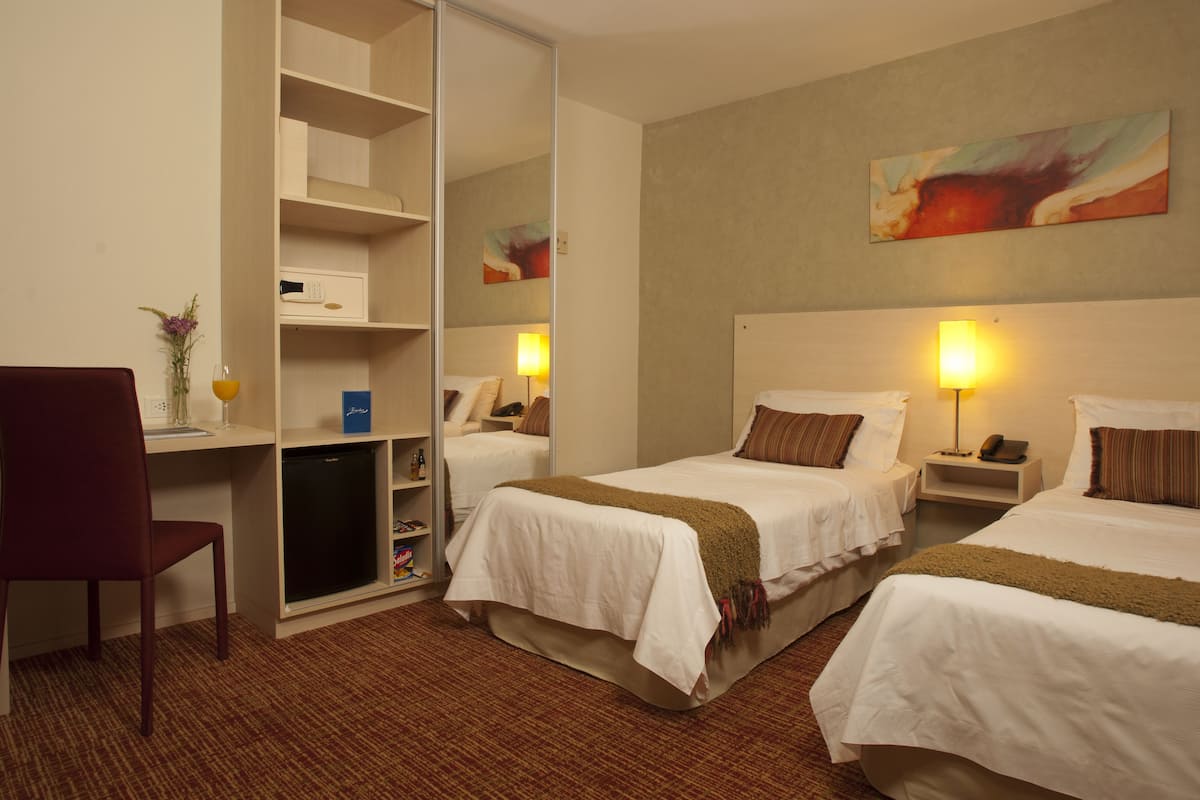 Ein Hotelzimmer mit zwei Betten, einem Schreibtisch, einem Stuhl, einem Kleiderschrank und einem Bild an der Wand.