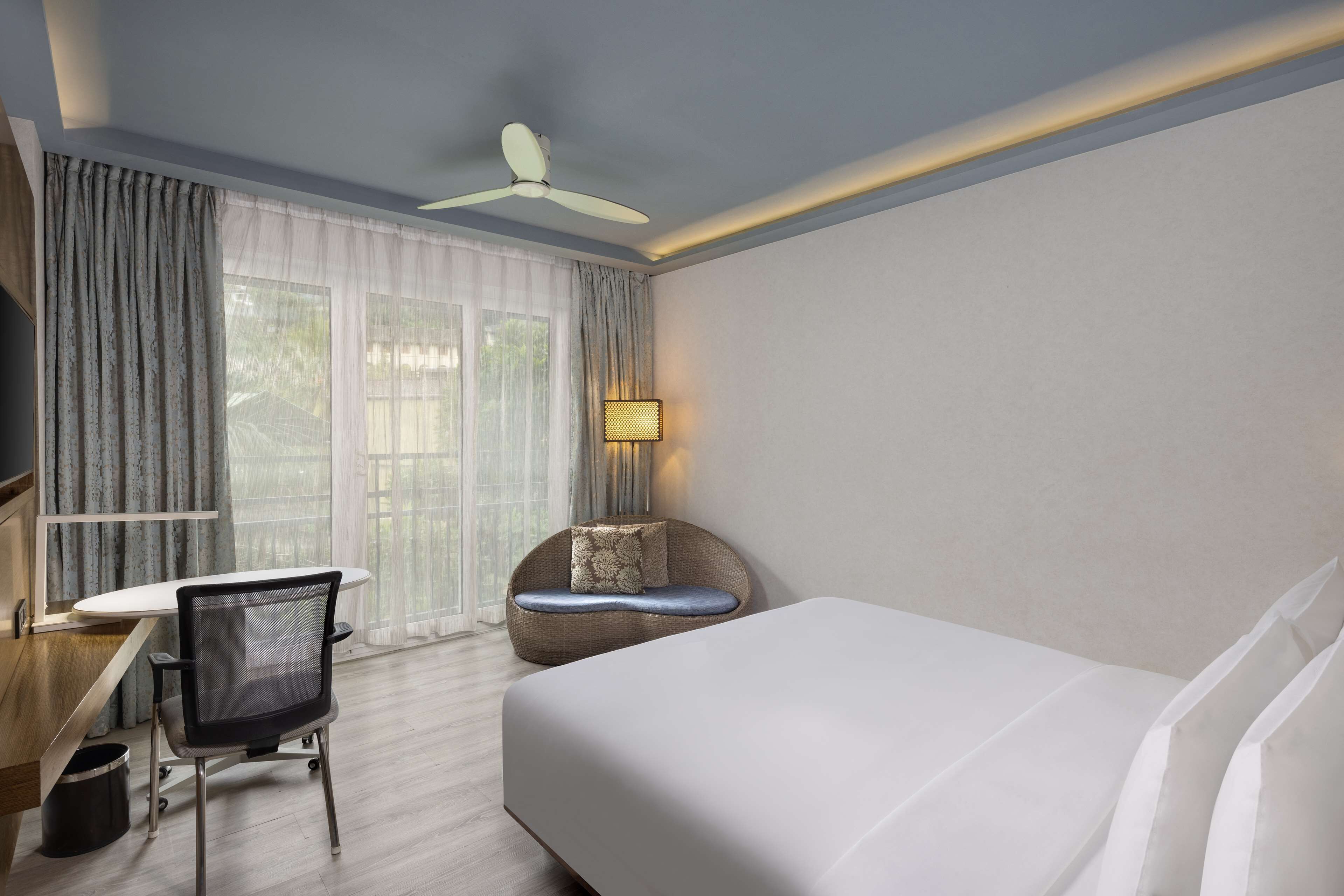 A hotel room with a bed, a desk, a chair, a ceiling fan, and a window with curtains.