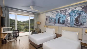 Two single beds with white linens, a flat-screen TV, and a desk with a chair, set against a backdrop of a large elephant mural.