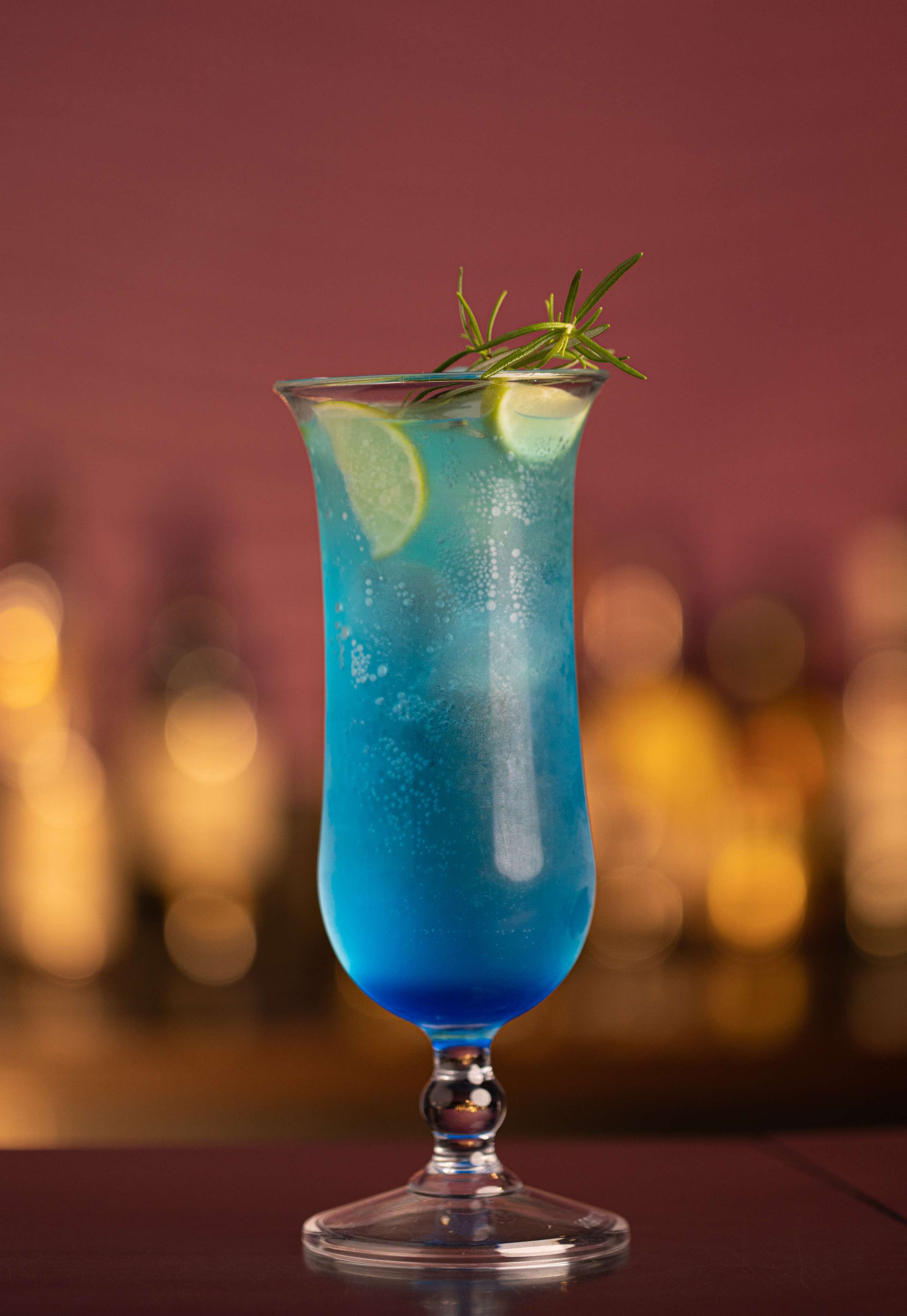 Un vaso alto con una bebida azul, adornado con una ramita de romero y una rodaja de lima, sobre un fondo desenfocado.