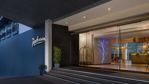 Una entrada de hotel moderna con fachada oscura, una escalera visible y un área de recepción con una exhibición digital de un árbol.