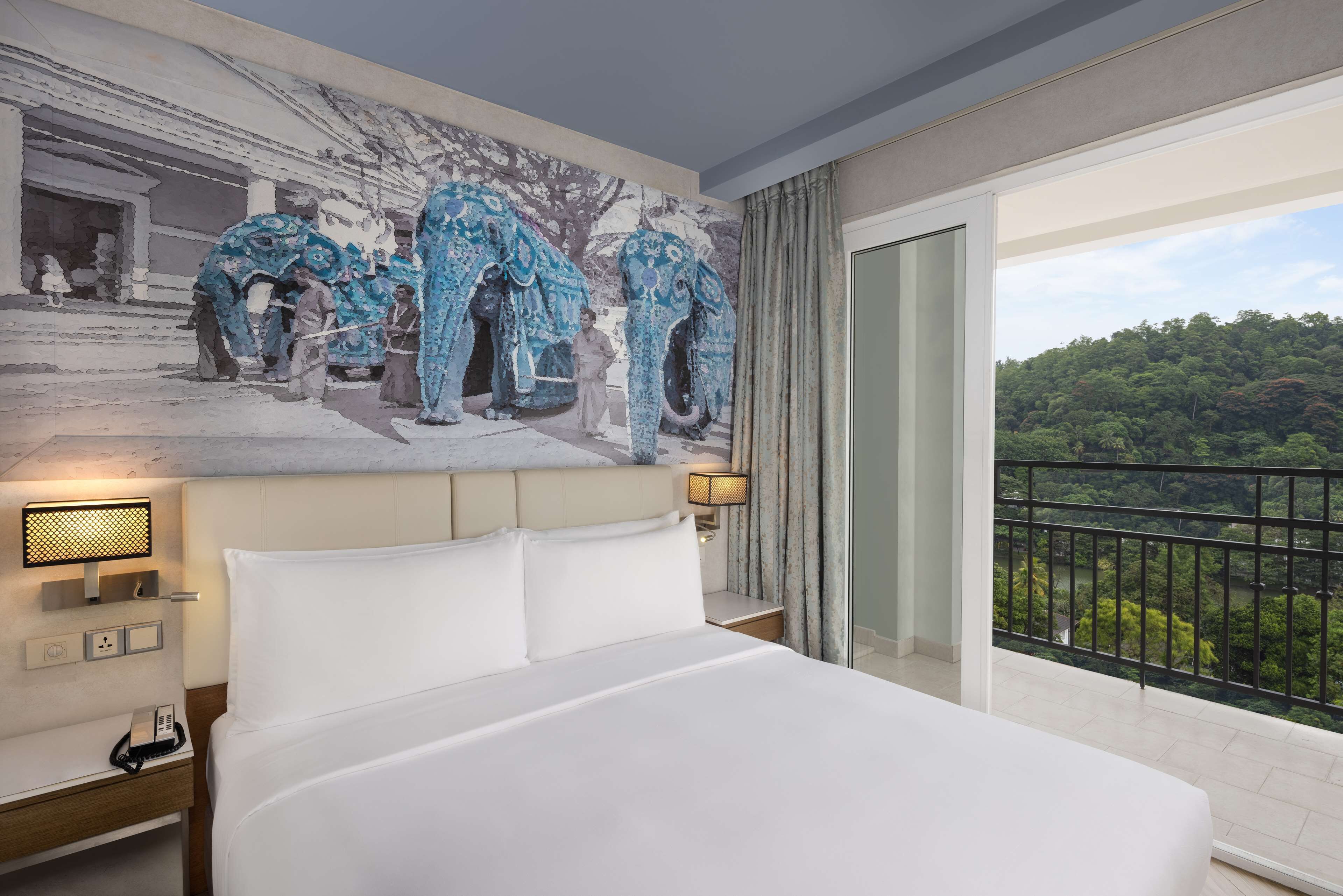 Habitación de hotel con una cama grande, una mesita de noche, una lámpara de pared y vistas a un área boscosa a través de un balcón.