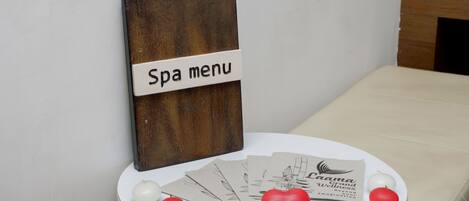 Een menukaart voor een spa op een houten bord, een witte tafel met afgeronde rand, en verschillende spa-kaarten en decoratieve ballen.