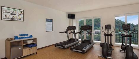 A gym with treadmills, elliptical machines, and a wall-mounted TV. There's a poster on the wall and a small cabinet with exercise equipment.