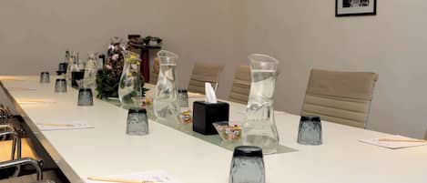 Un salón de conferencias con una larga mesa, sillas y diversos objetos como jarras de vidrio, una caja de pañuelos y un cuadro en la pared.