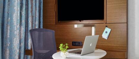 Una habitación moderna con televisor montado en la pared, una laptop sobre una mesita, una silla y una planta.