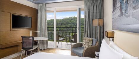 Une chambre d’hôtel avec un grand lit, un téléviseur fixé au mur, un balcon avec une table et des chaises, et un tableau accroché au mur.