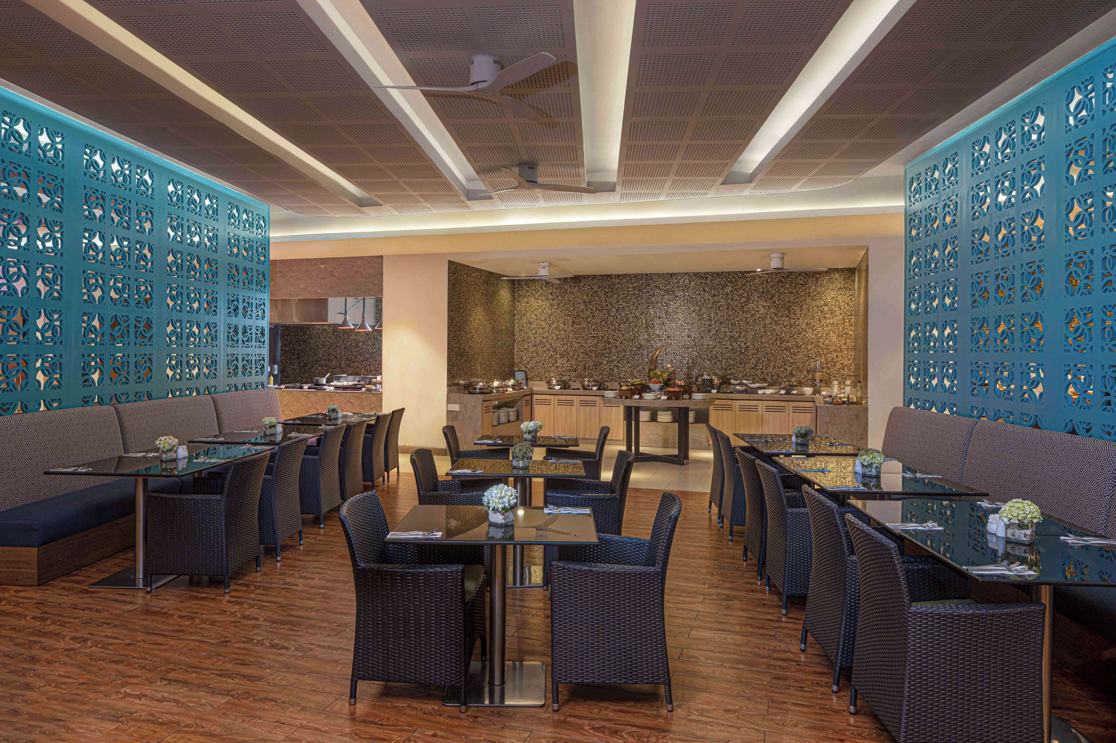 A restaurant with multiple dining tables and chairs, a kitchen area in the background, and decorative blue panels on the walls.