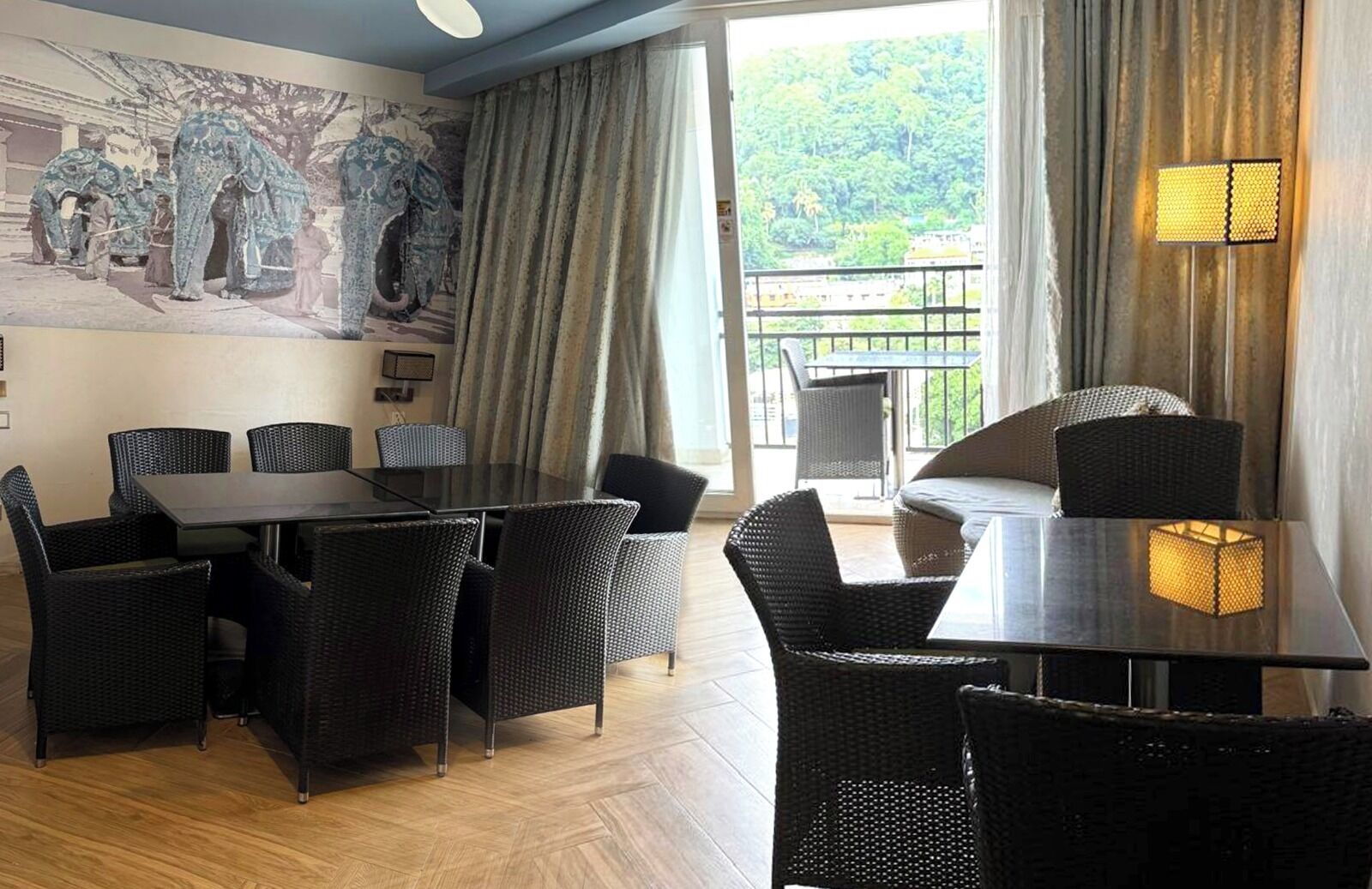 A dining area with a large table and chairs, a balcony with a view of greenery, and a wall with a mural.