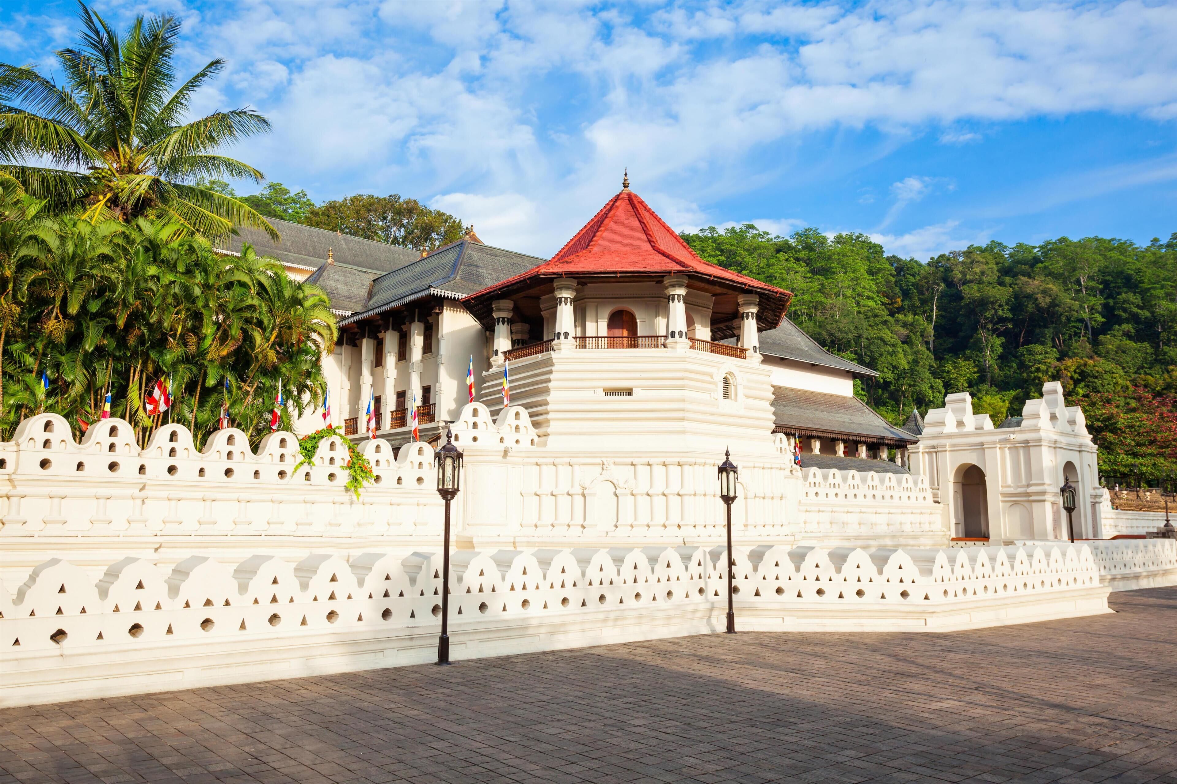 A traditional white building with a red roof, surrounded by a white wall with decorative elements, set against a backdrop of lush greenery and a clear sky.