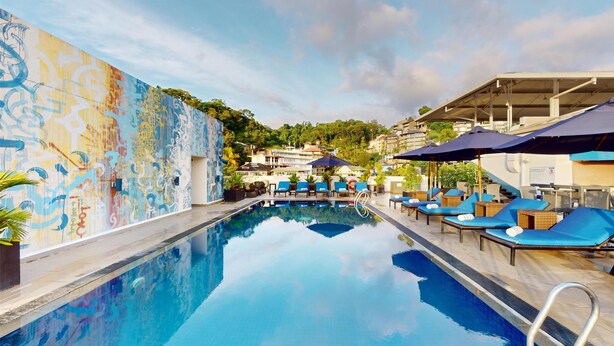 Una piscina con sillones de descanso y sombrillas, rodeada por un mural colorido y vegetación.