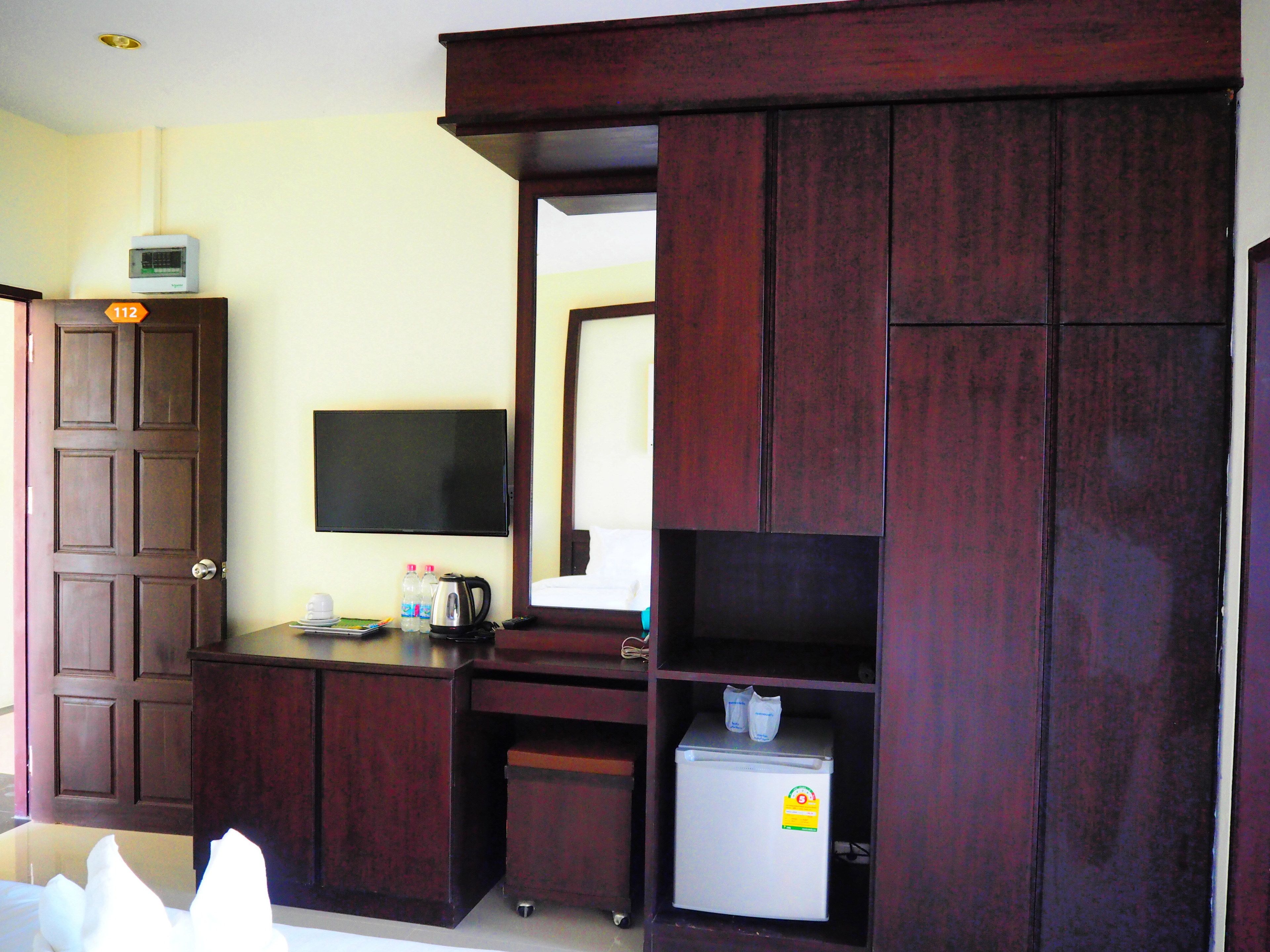A compact hotel room with a wooden wardrobe, a small refrigerator, a flat-screen TV, and a desk with a kettle and bottles.