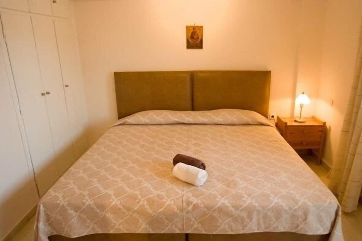 A neatly made bed with a patterned bedspread, a pillow, and a rolled towel on the bed. There is a small bedside table with a lamp and a wall-mounted picture above the bed.