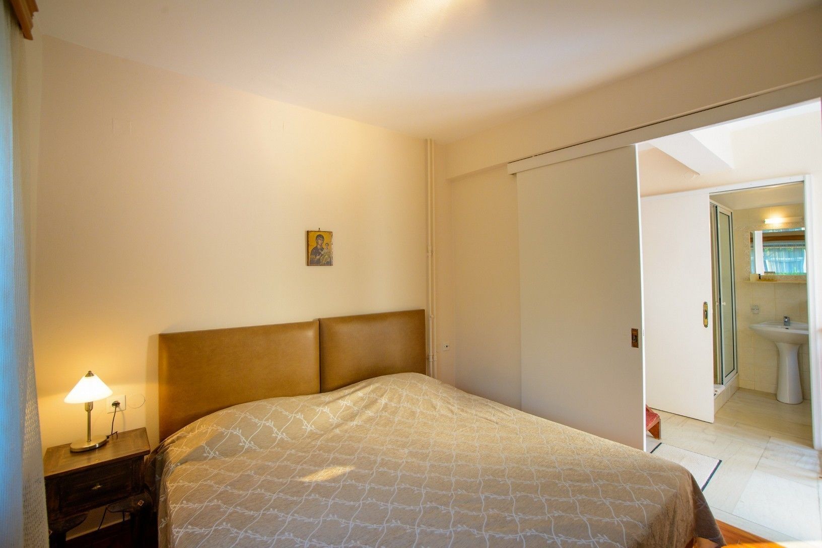 Une chambre à coucher comprenant un lit, une table de chevet, une lampe, un tableau accroché au mur et une porte donnant sur une salle de bain.
