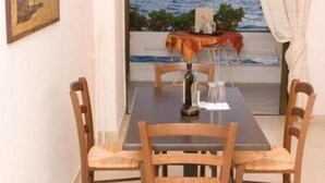 A dining area with a table and four chairs, a painting on the wall, and a view of the sea through a window with curtains.