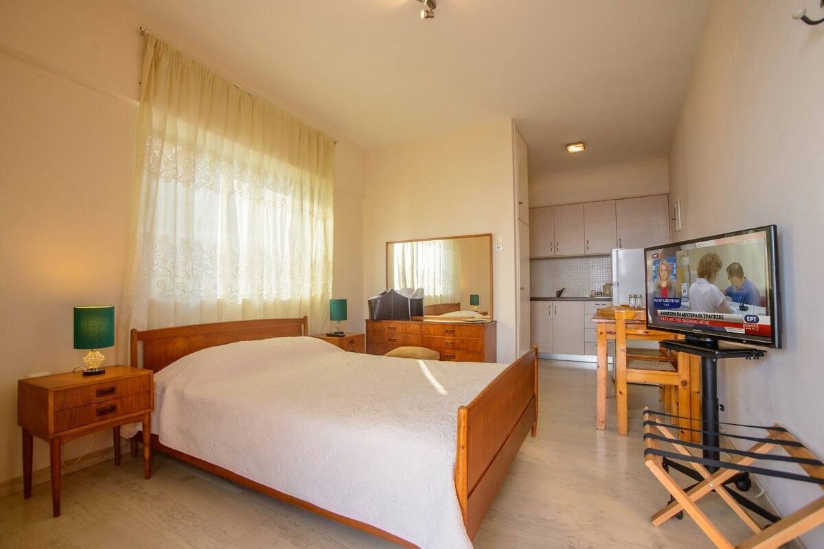 A hotel room with a bed, bedside tables, a desk with a chair, a television, and a kitchen area.