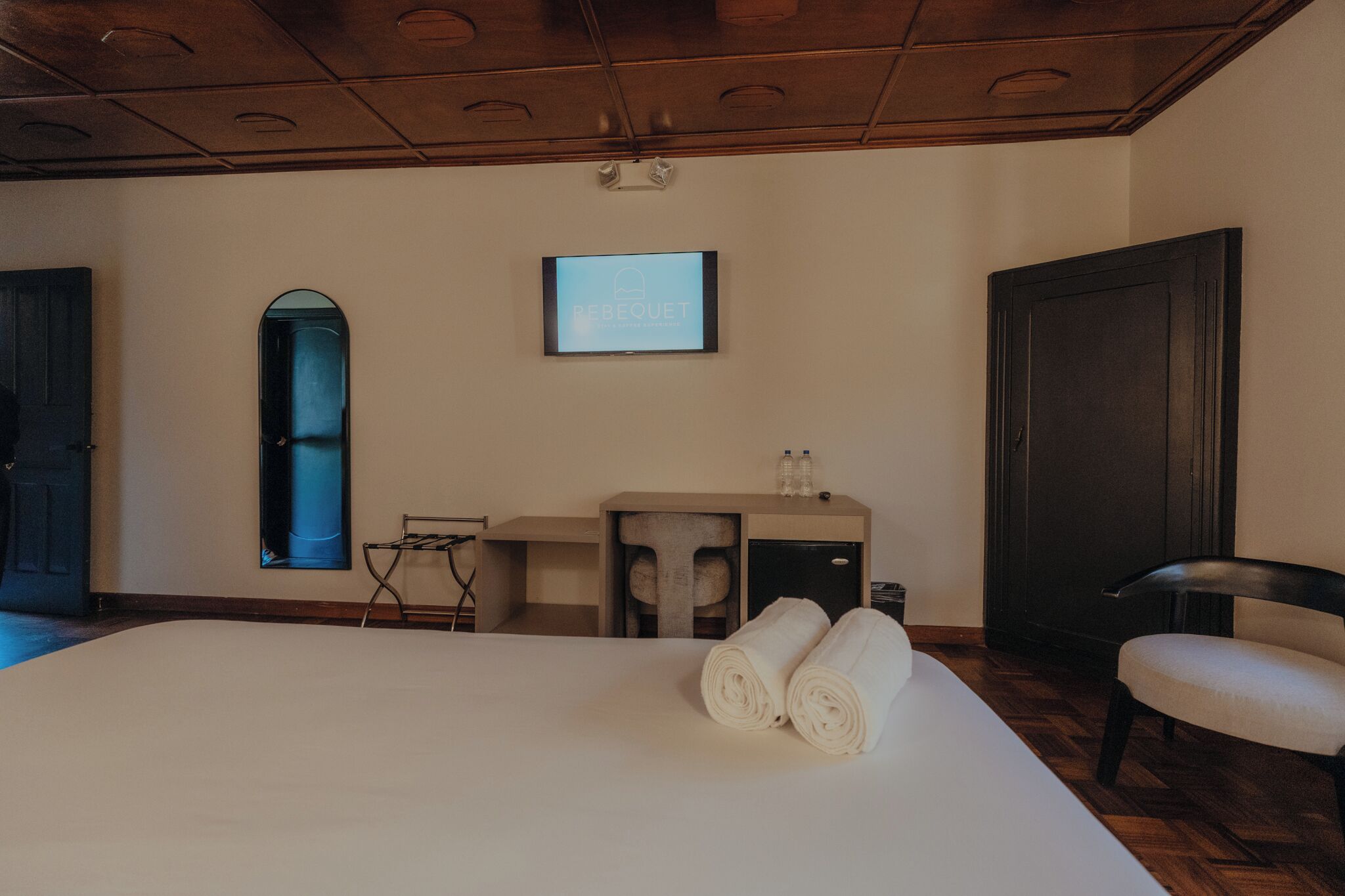 Une chambre d’hôtel avec un grand lit, un bureau avec une chaise, une petite table, un miroir et un écran de télévision affichant le mot « REBEQUET ».