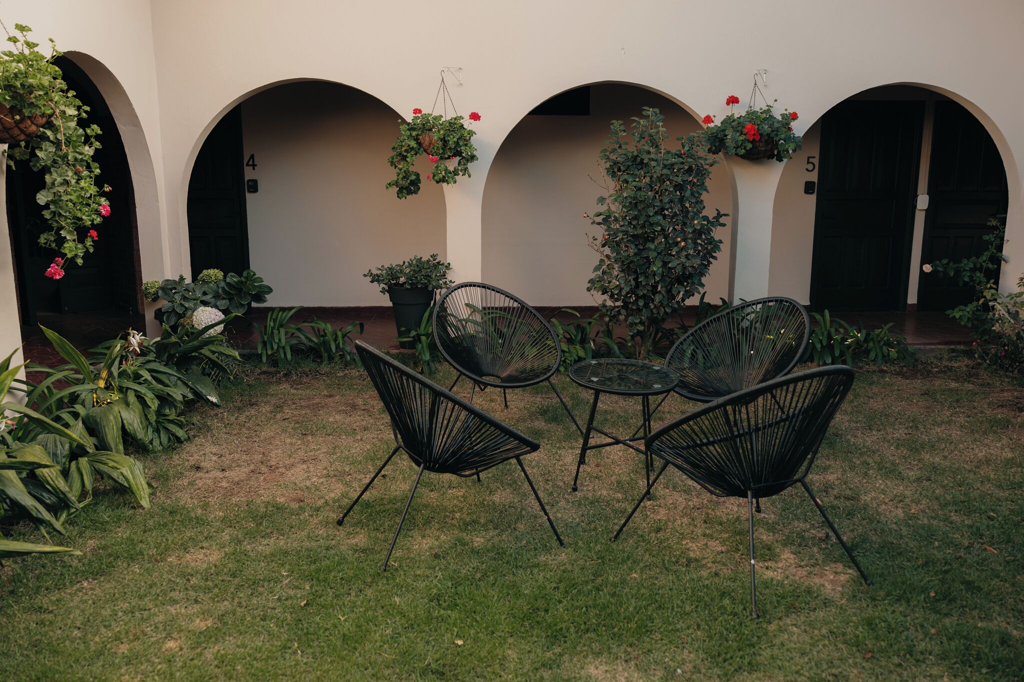 Un área de estar al aire libre con sillas negras y una pequeña mesa, rodeada de plantas en macetas y cestas colgantes de flores.