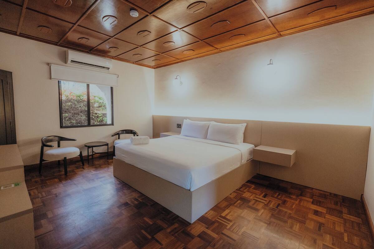 Un dormitorio con una cama grande, una ventana, una silla, una mesita pequeña y una mesita de noche.