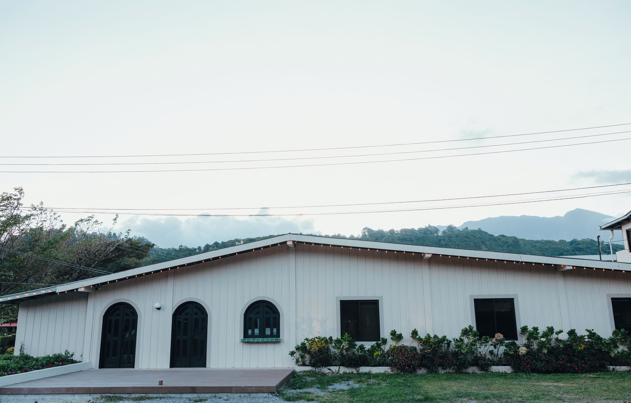 Un edificio blanco con ventanas arqueadas y techo plano, rodeado de vegetación y montañas al fondo.