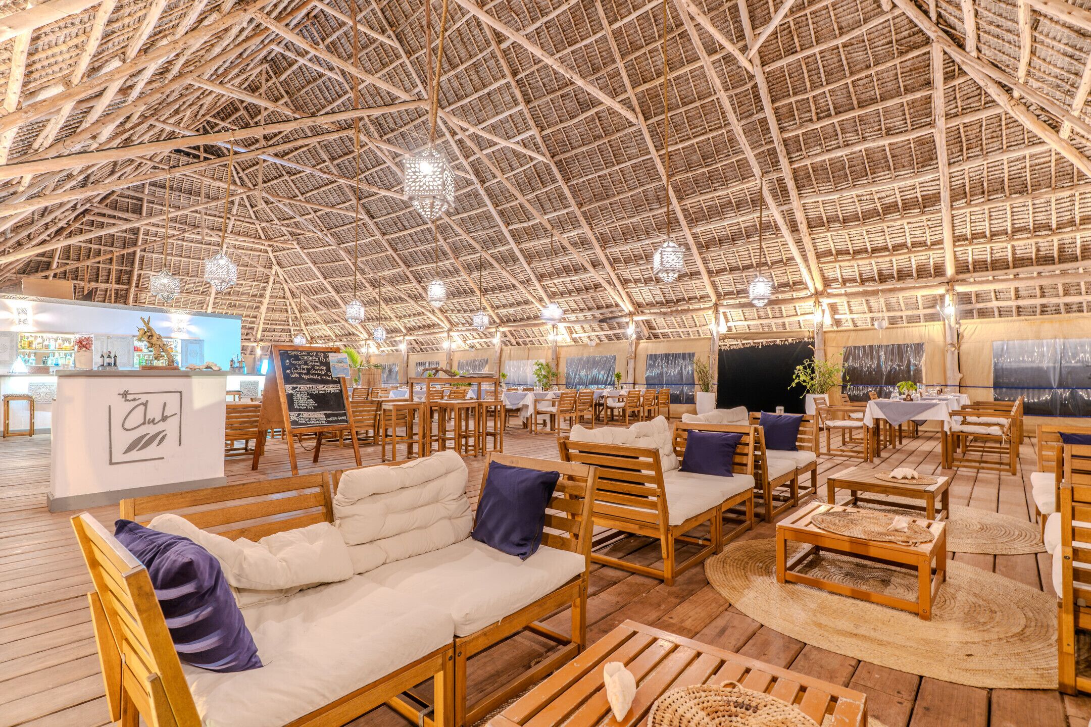 An indoor venue with a high thatched roof, wooden furniture, and a bar area.