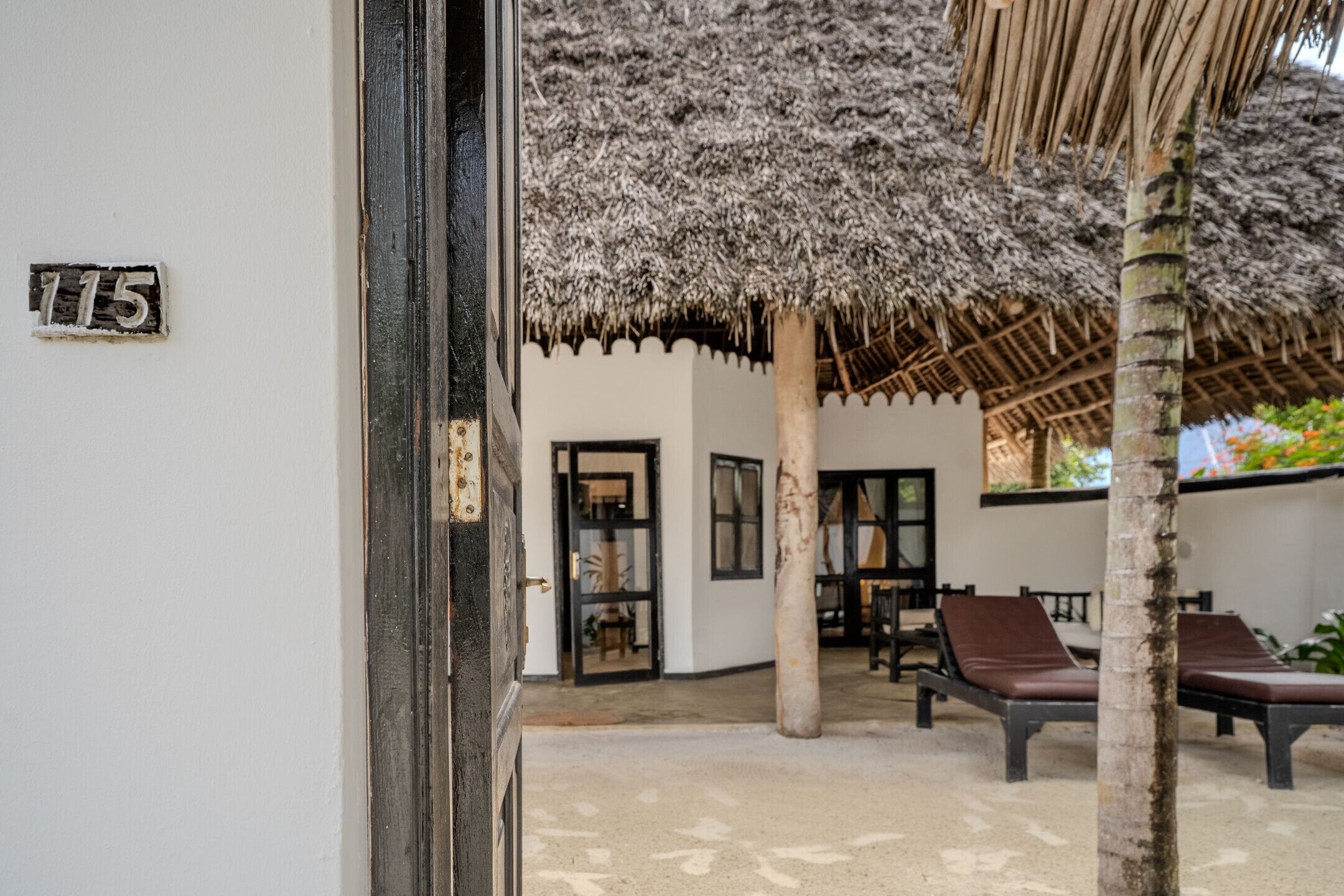 L’entrée d’un hôtel, avec une enseigne portant le numéro « 115 », un espace extérieur abrité avec des chaises, et une structure dotée d’un toit de chaume.