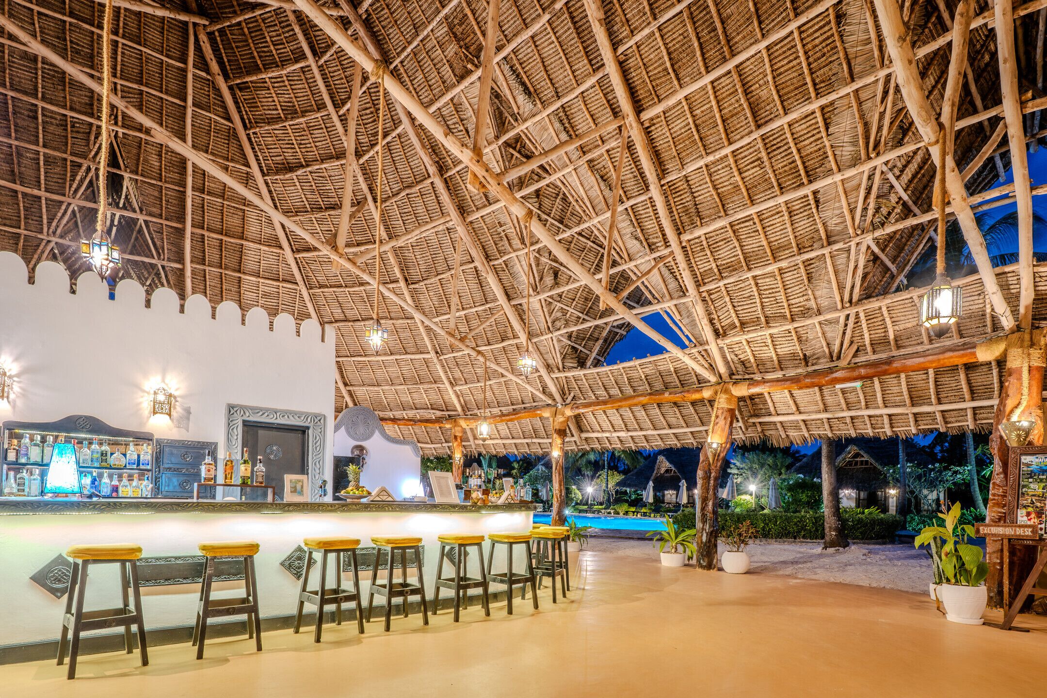 Un espace bar extérieur doté d’un toit de chaume, de tabourets de bar et d’une piscine en arrière-plan.