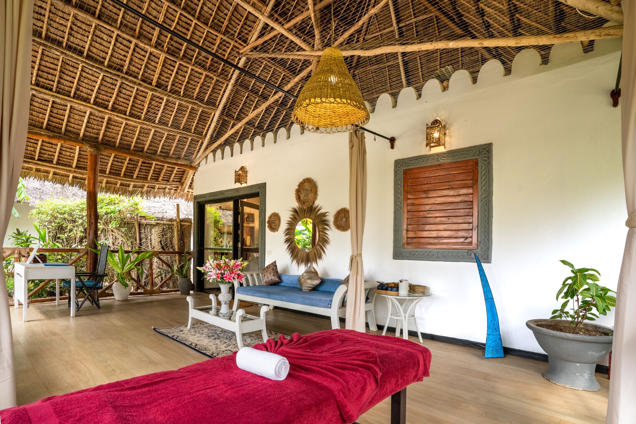 A room with a thatched roof, a massage table with a red towel, a blue sofa, and a wooden window shutter.