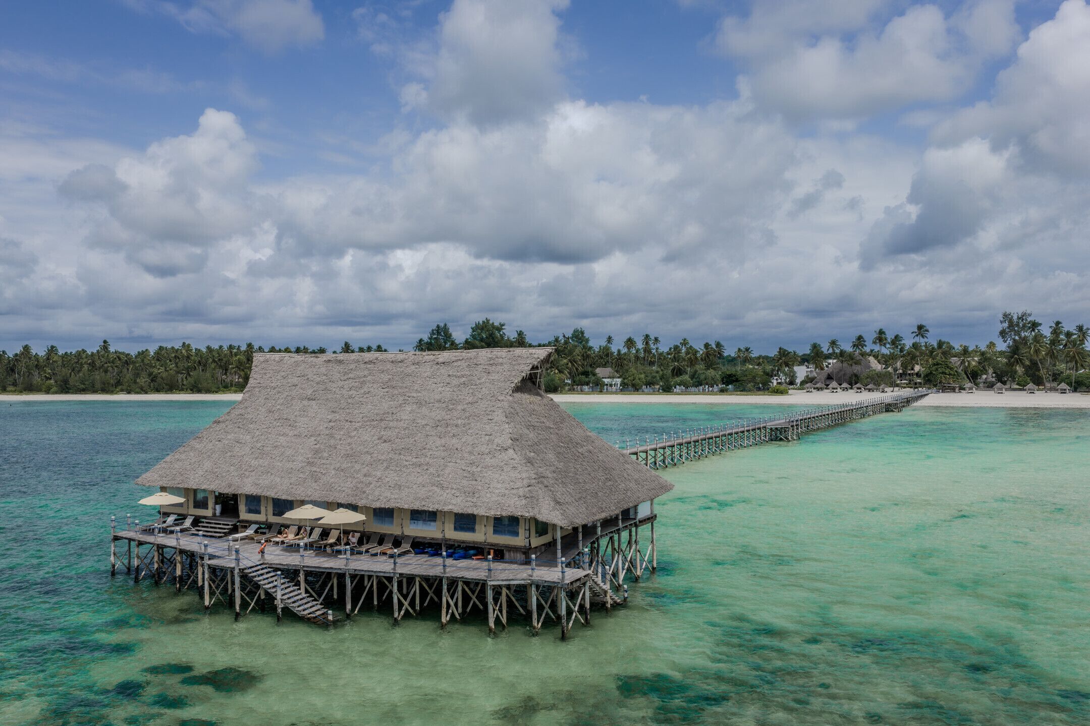 An overwater bungalow with a thatched roof, multiple lounge chairs, and a wooden deck.