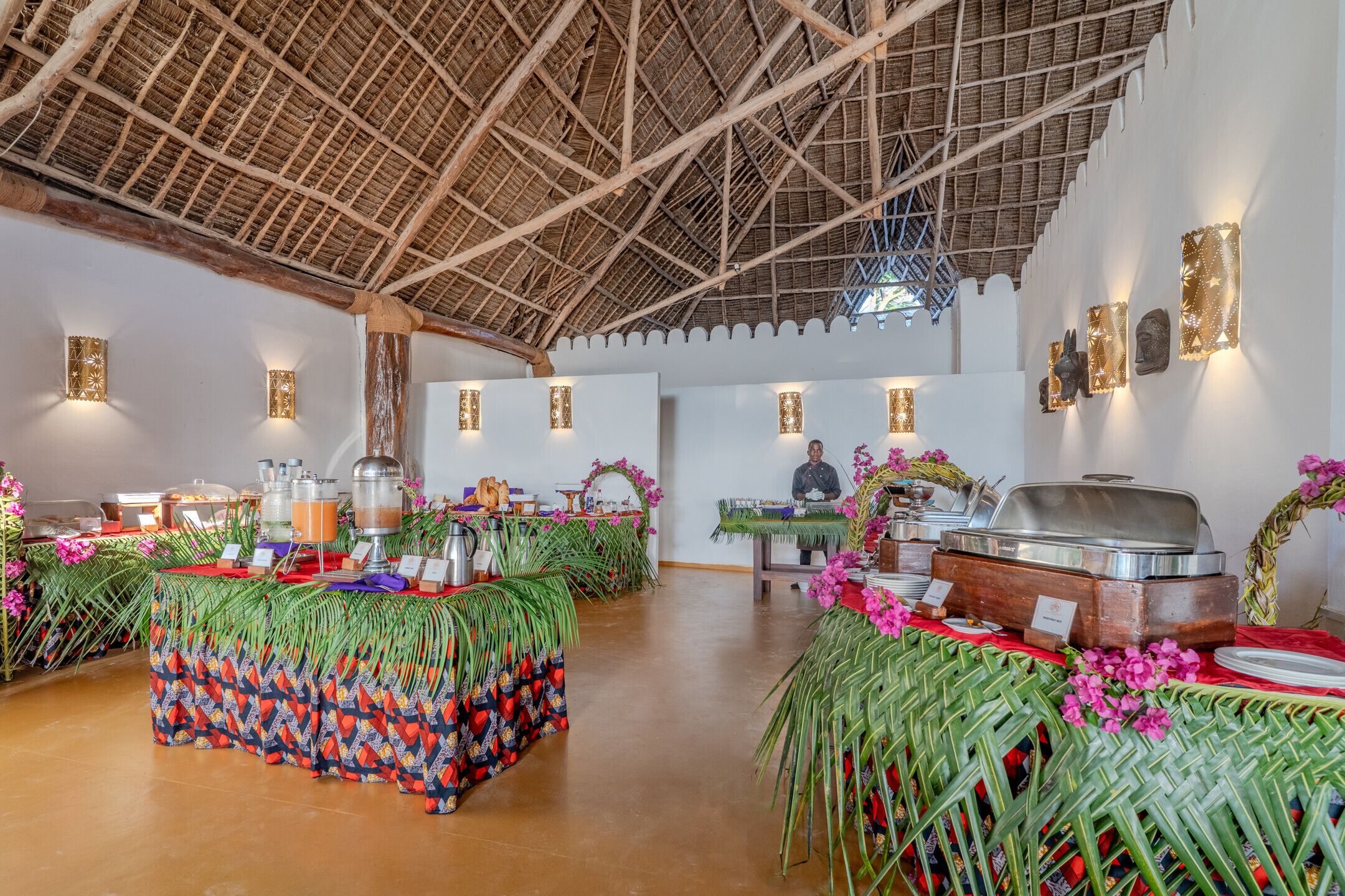 A spacious indoor area with a high ceiling and thatched roof structure. There are buffet tables with food and drink, and decorative elements like flowers and wall art.