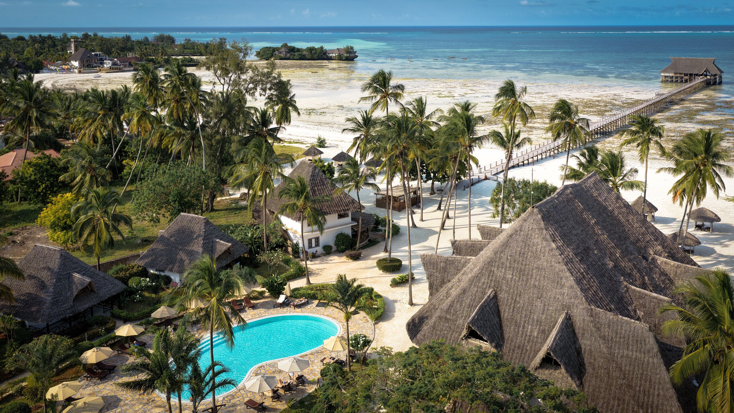 A resort with a swimming pool, thatched-roof structures, palm trees, and a beach view.