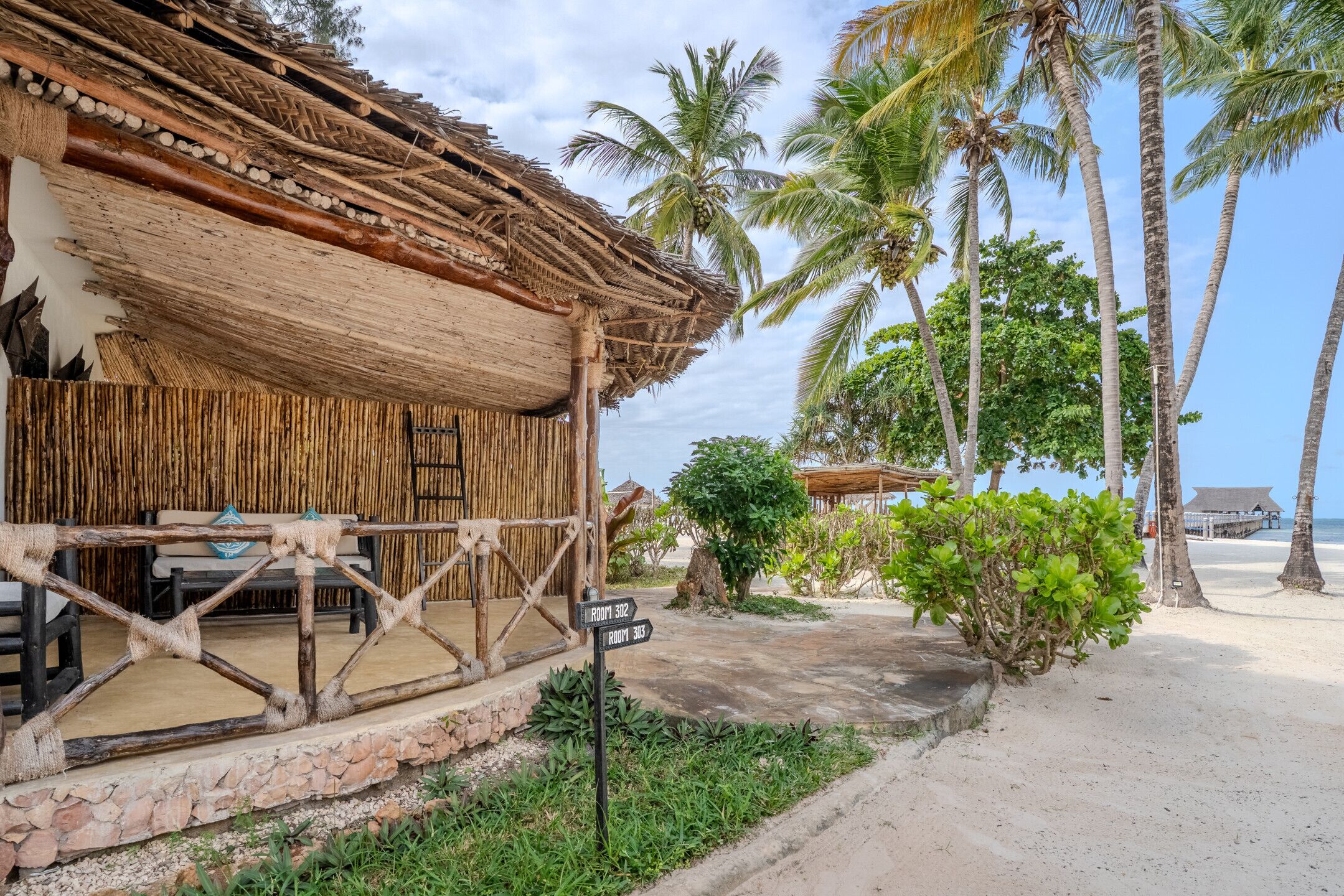 A thatched-roof building with a wooden deck, palm trees, and a sign indicating rooms 302 and 303.